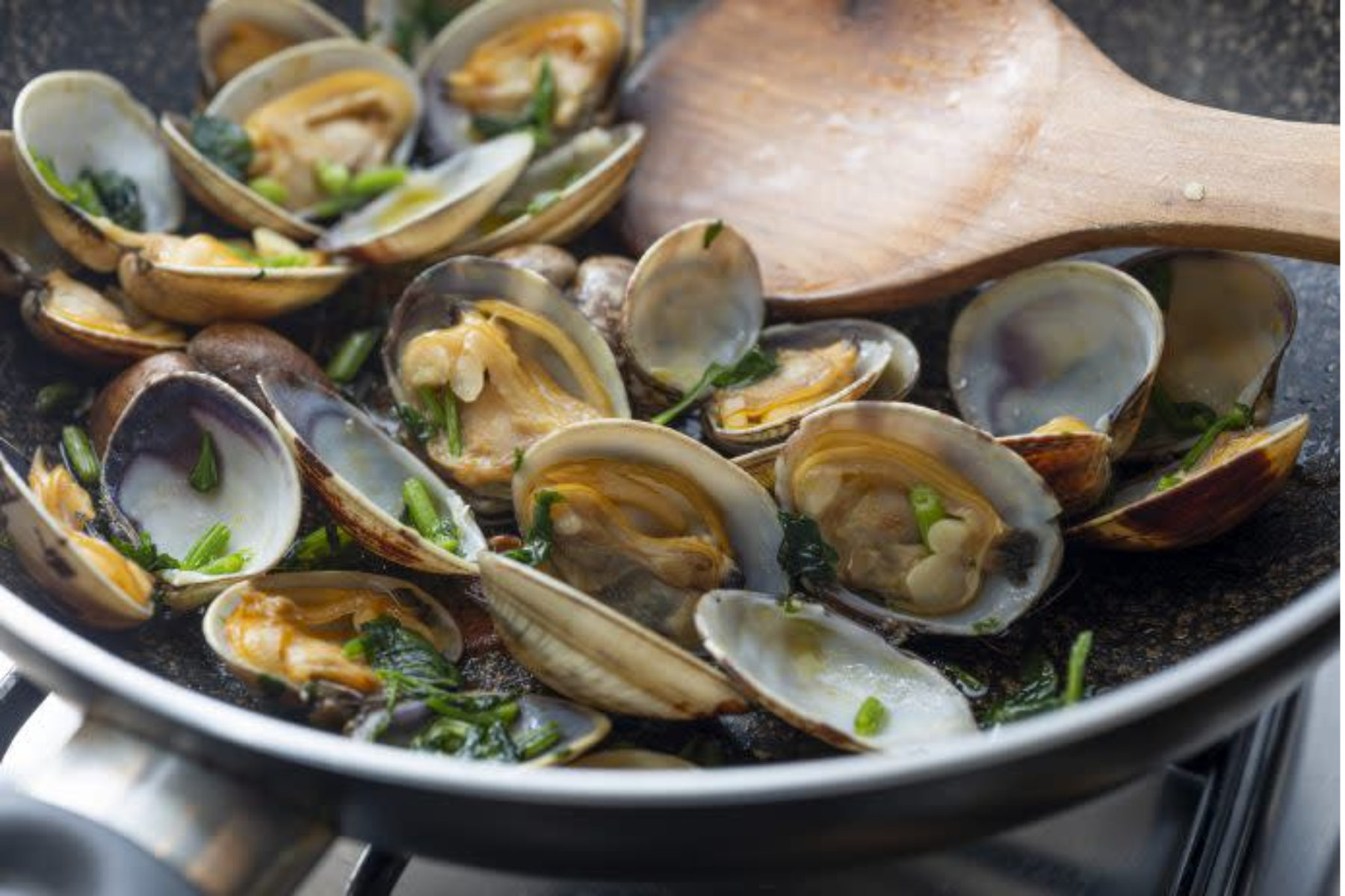 Tons of clams being cooked in a frying pan with a wooden ladle