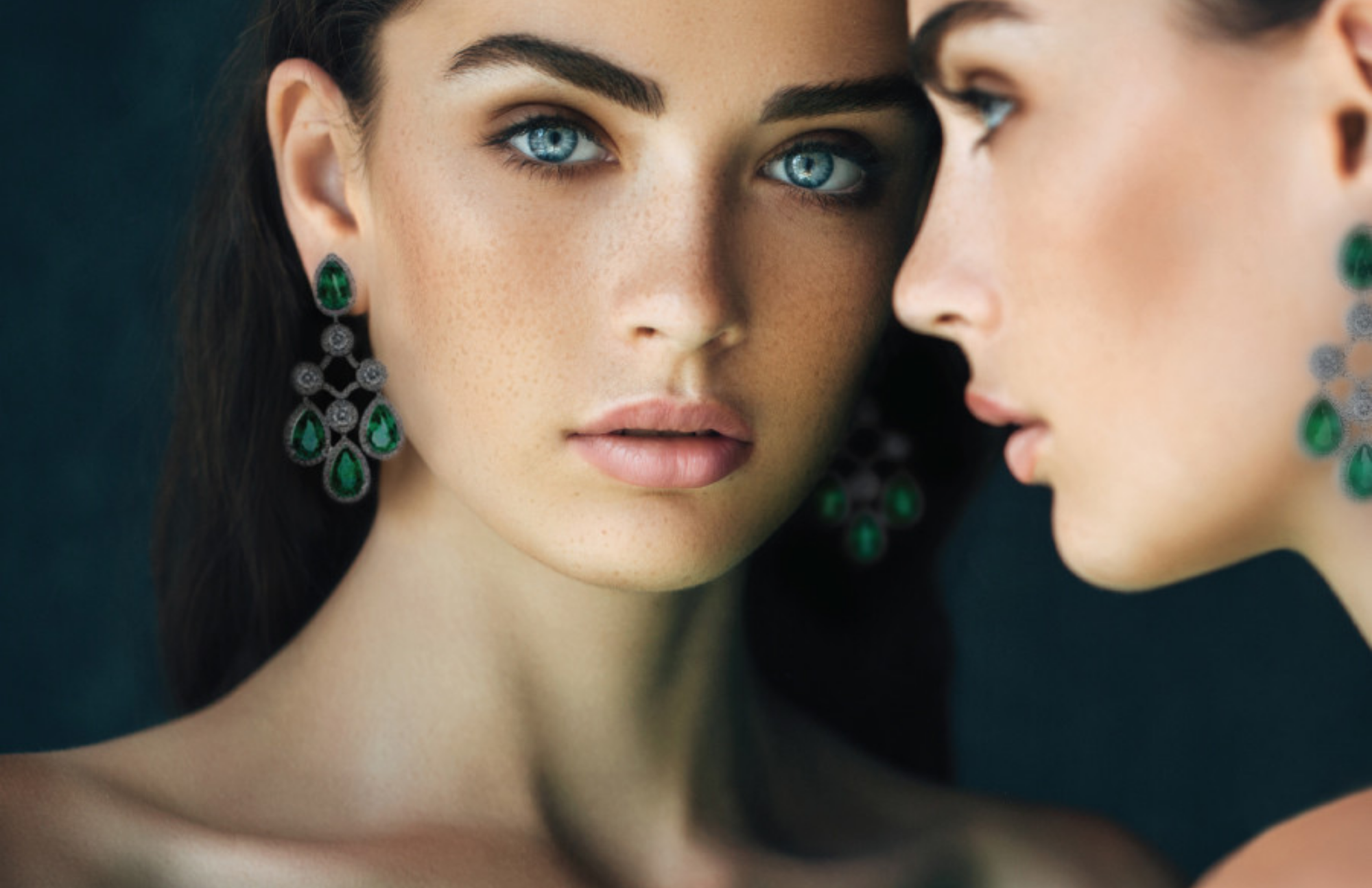 A woman's reflection in a mirror looking directly at the camera while wearing an emerald earring