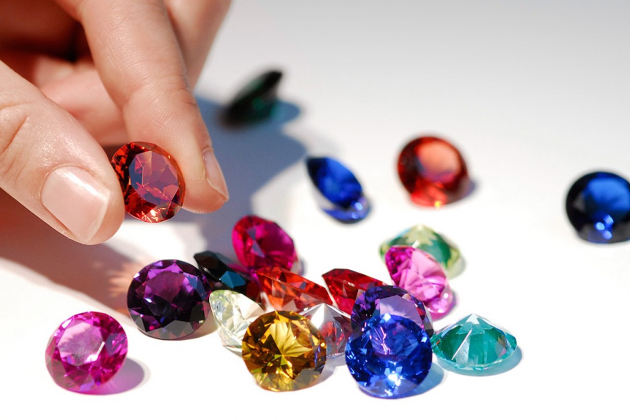 Blue, transparent, green, pink, and yellow small gemstones with a hand holding a red gemstone