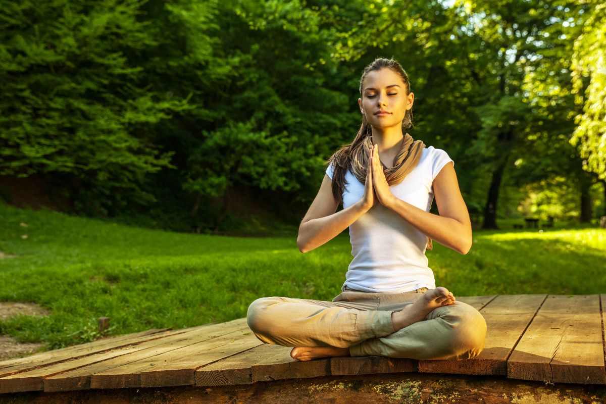 A woman is meditating while sitting on a bench in the woods, surrounded by trees