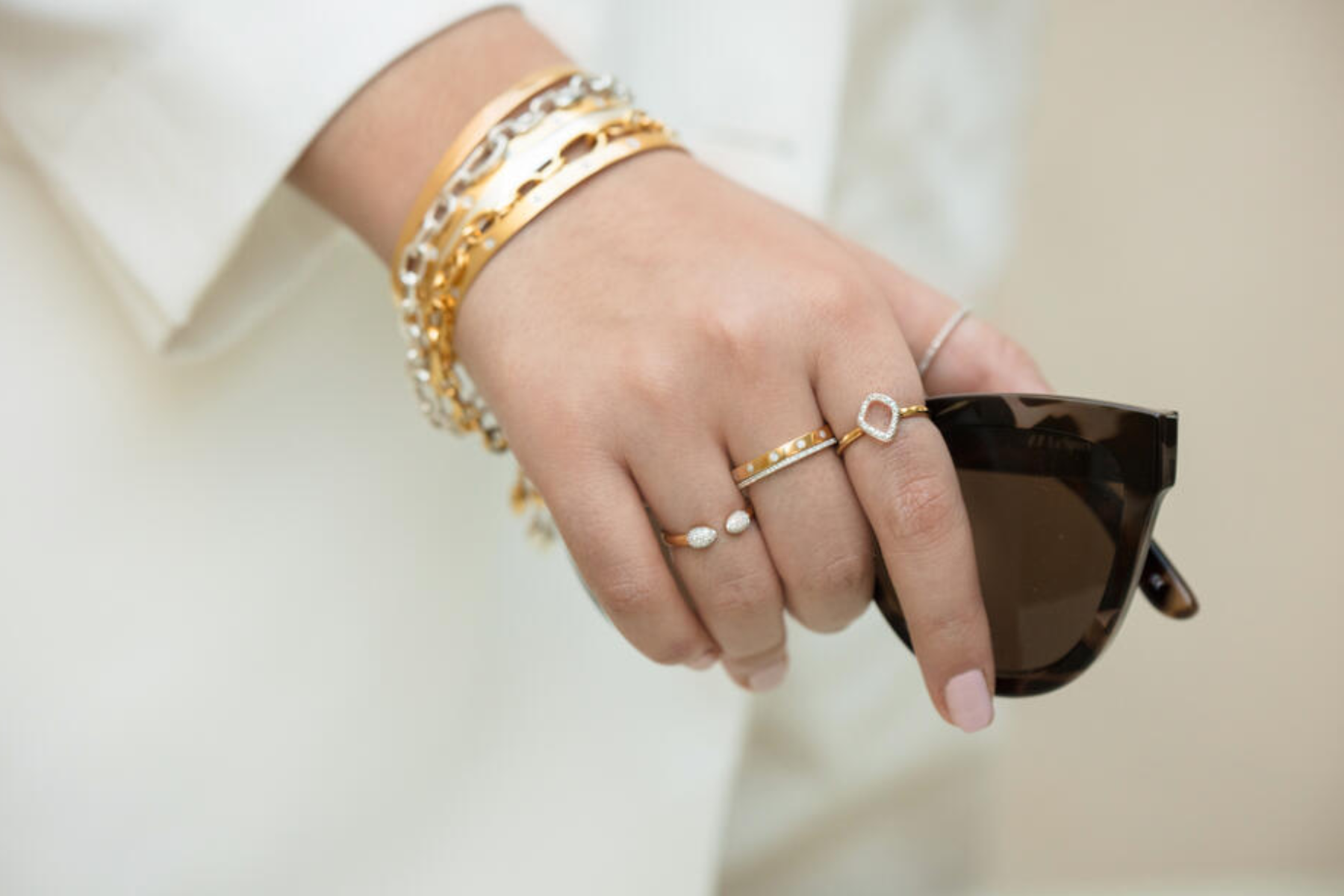 A woman in white wearing gold bracelets and rings and holding a sunglass
