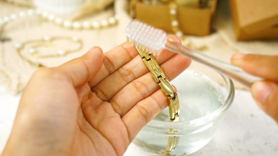 A woman's hand cleaning a gold bracelet with a soft brush and water