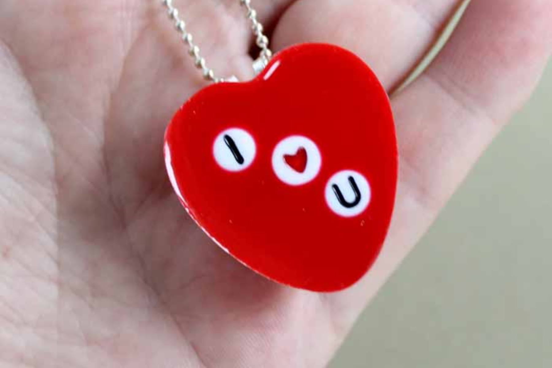 A woman's hand holding a red heart-shaped pendant with the words "I <3 U" engraved on it