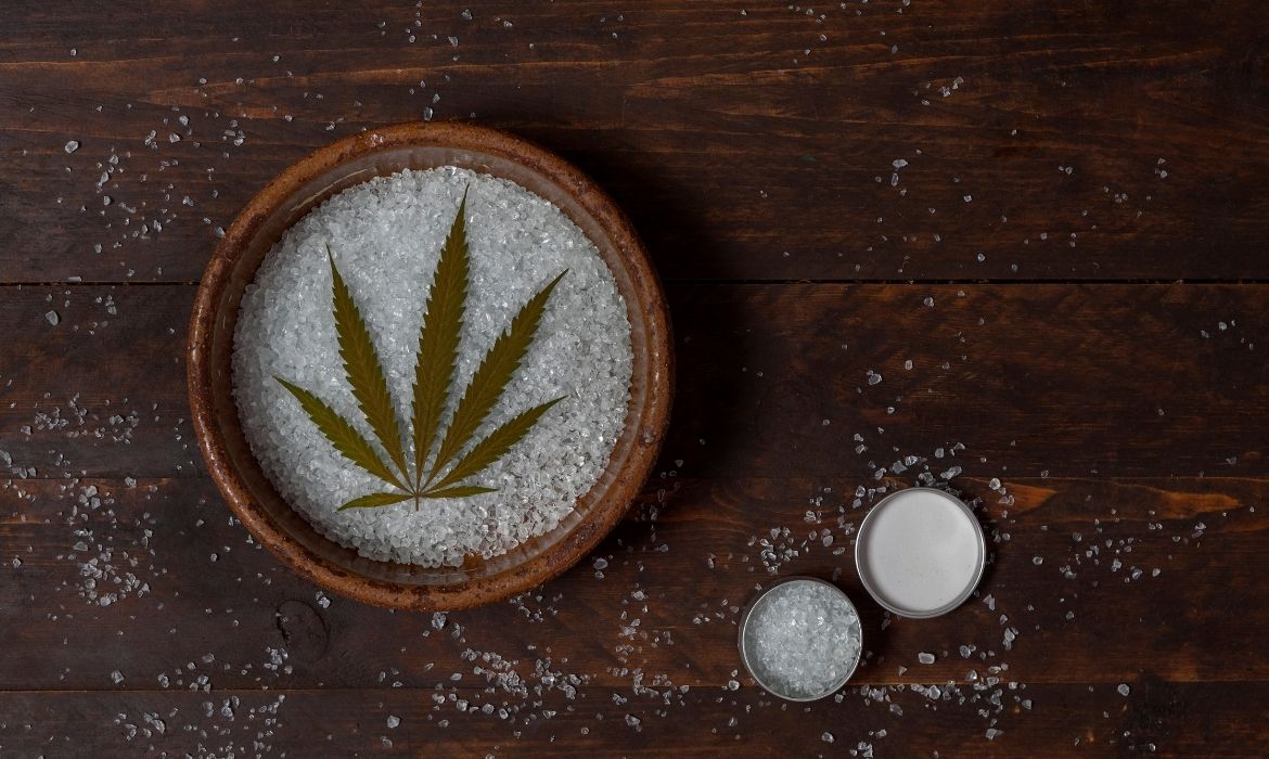 A wooden bowl filled with CBD crystals with a cannabis leaf in it beside an opened small can filled with CBD crystals with the cover by its side