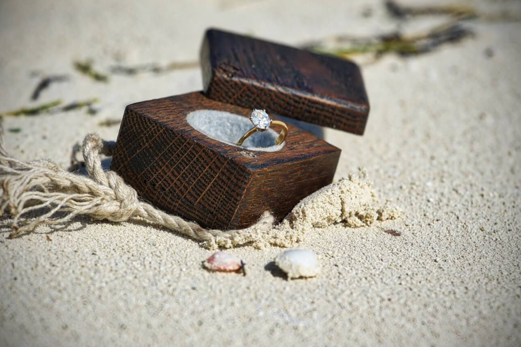 At the beach's sand, an oval-cut diamond engagement ring is displayed in a wooden box