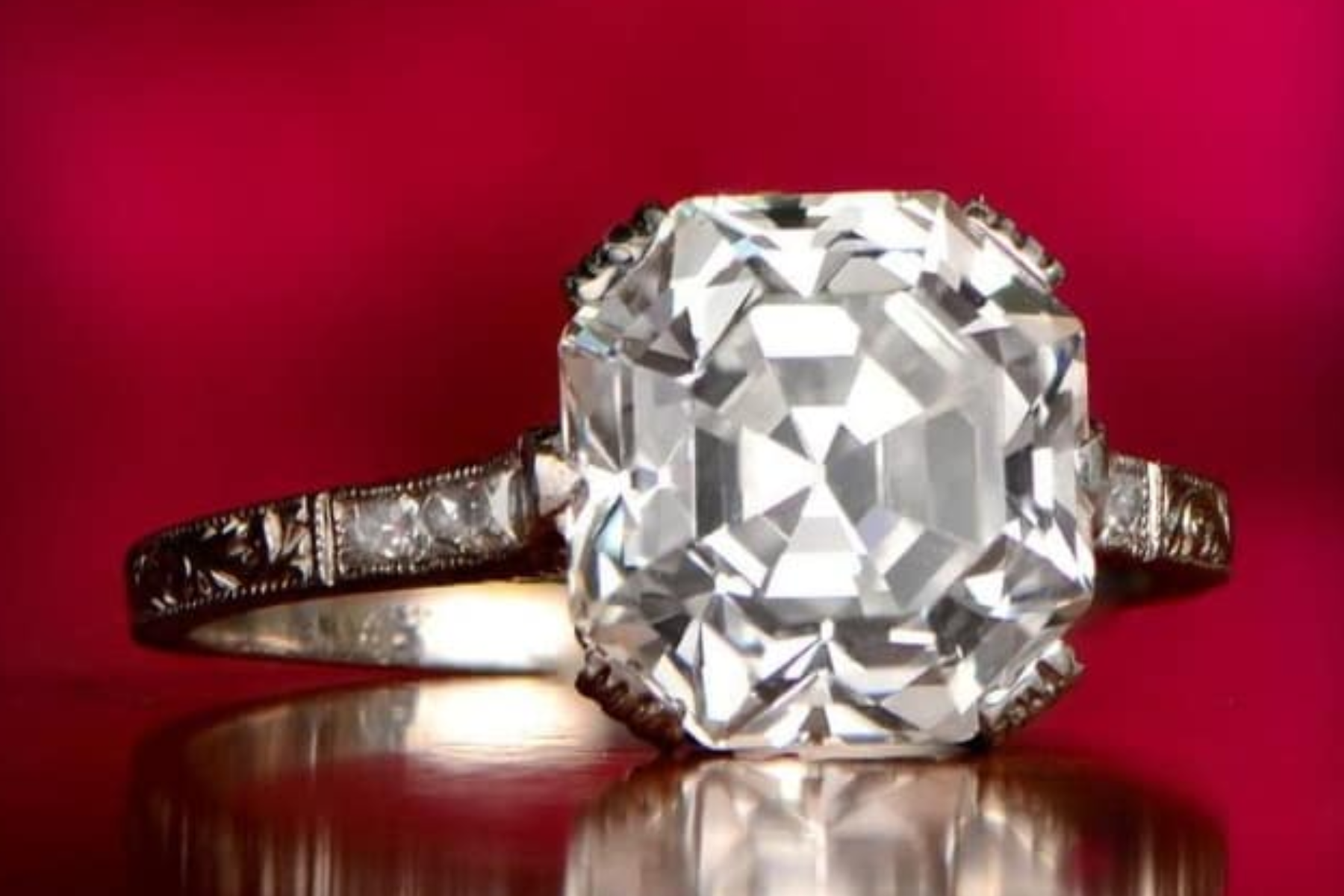 A close-up photograph of an engagement ring featuring an Asscher cut diamond set in a band. The ring is placed on a dark red background
