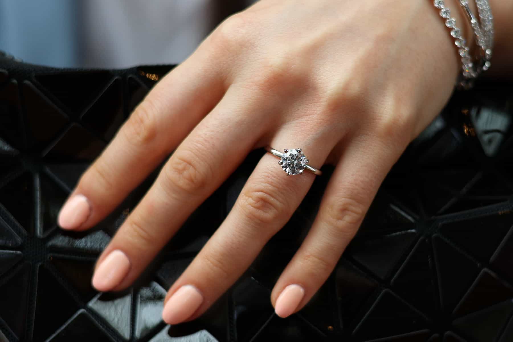 A close-up view of a woman's hand holding a black pouch. The hand is adorned with a round-cut engagement ring
