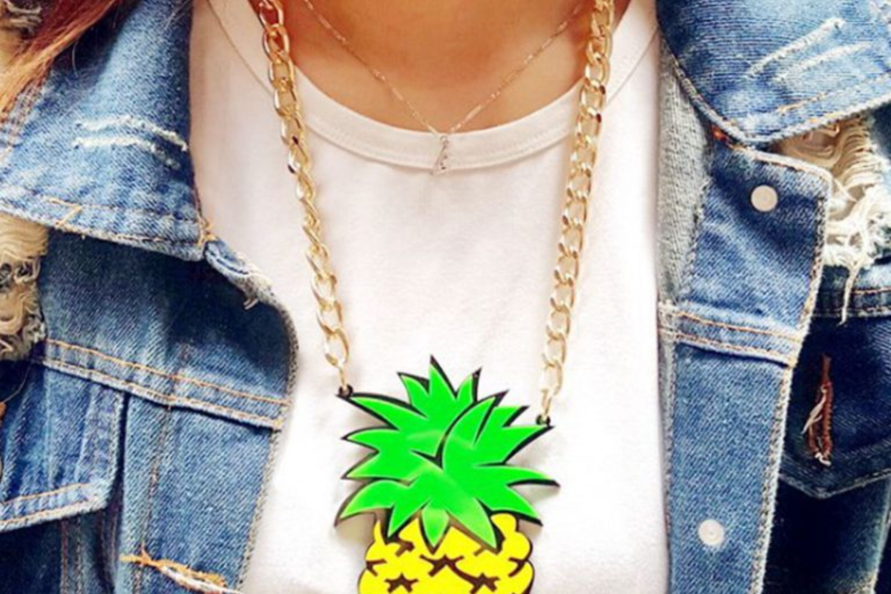 A close-up image of a woman's torso wearing a blue jean jacket. The woman is wearing a large pineapple pendant on a gold chain around her neck