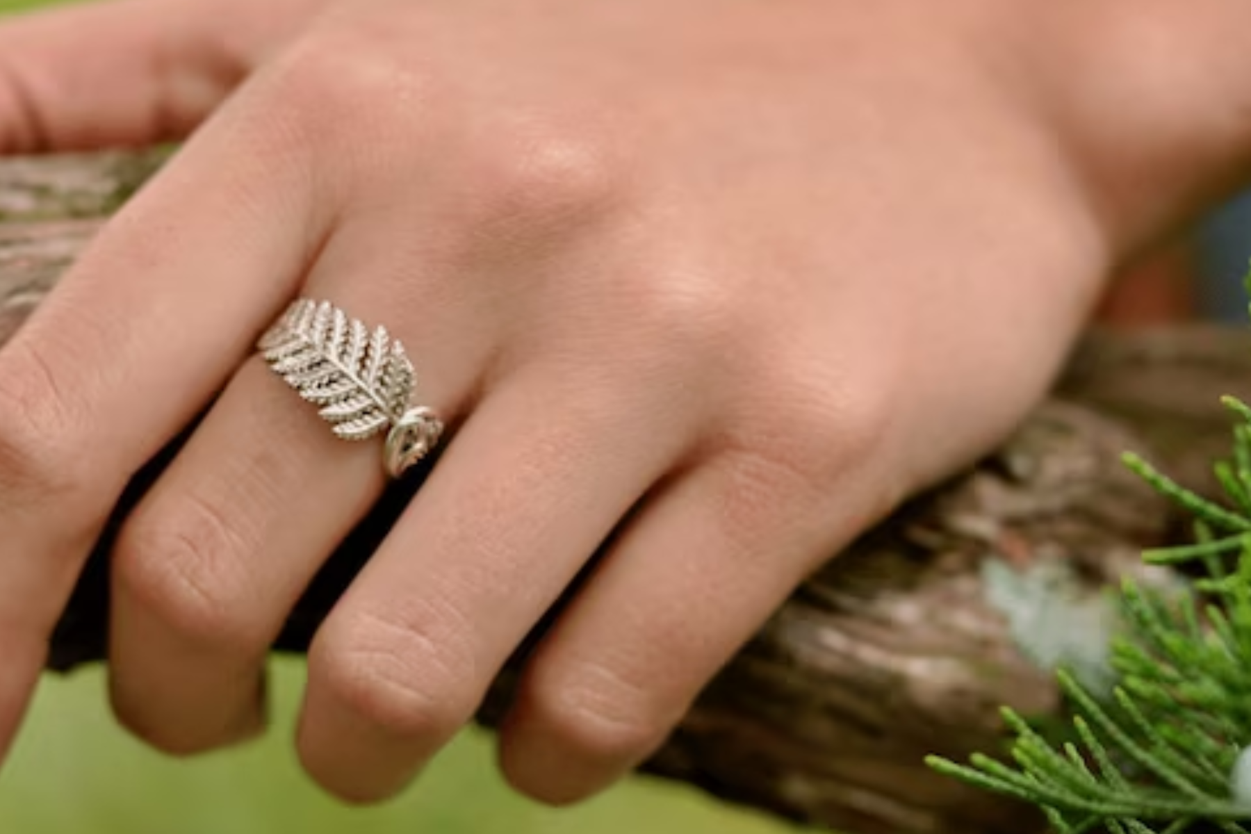 A close-up of a woman's hand holding a branch of a tree. The hand is adorned with a leaf ring