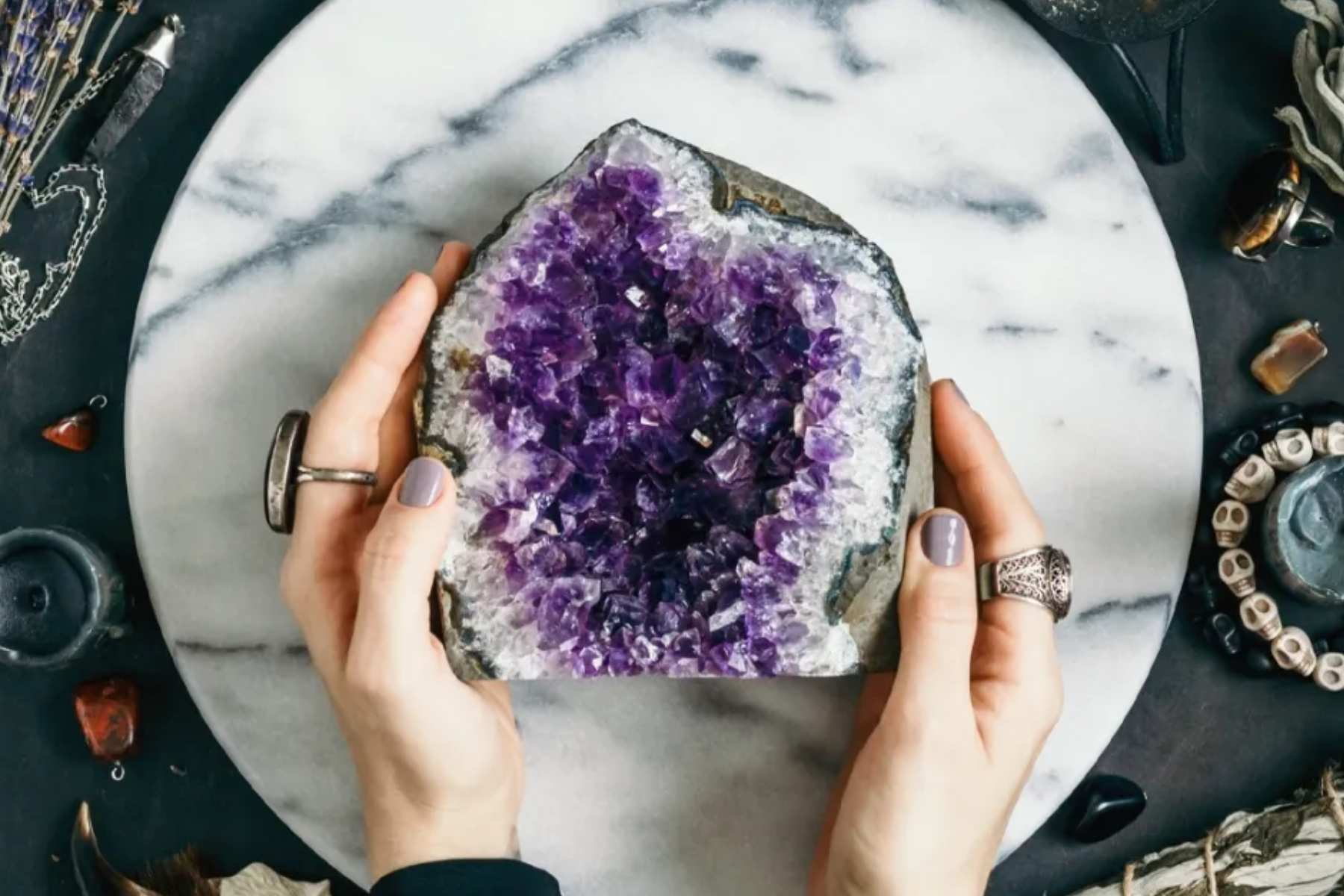 A photograph of a woman's hand grasping a large, unpolished amethyst crystal resting on a tabletop