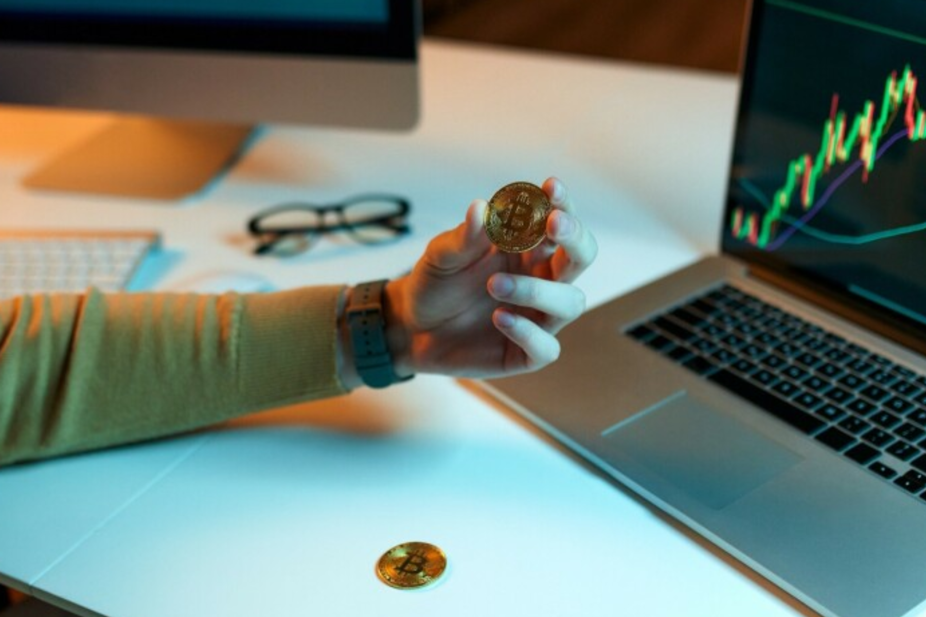 A Bitcoin is held by a hand in front of a laptop