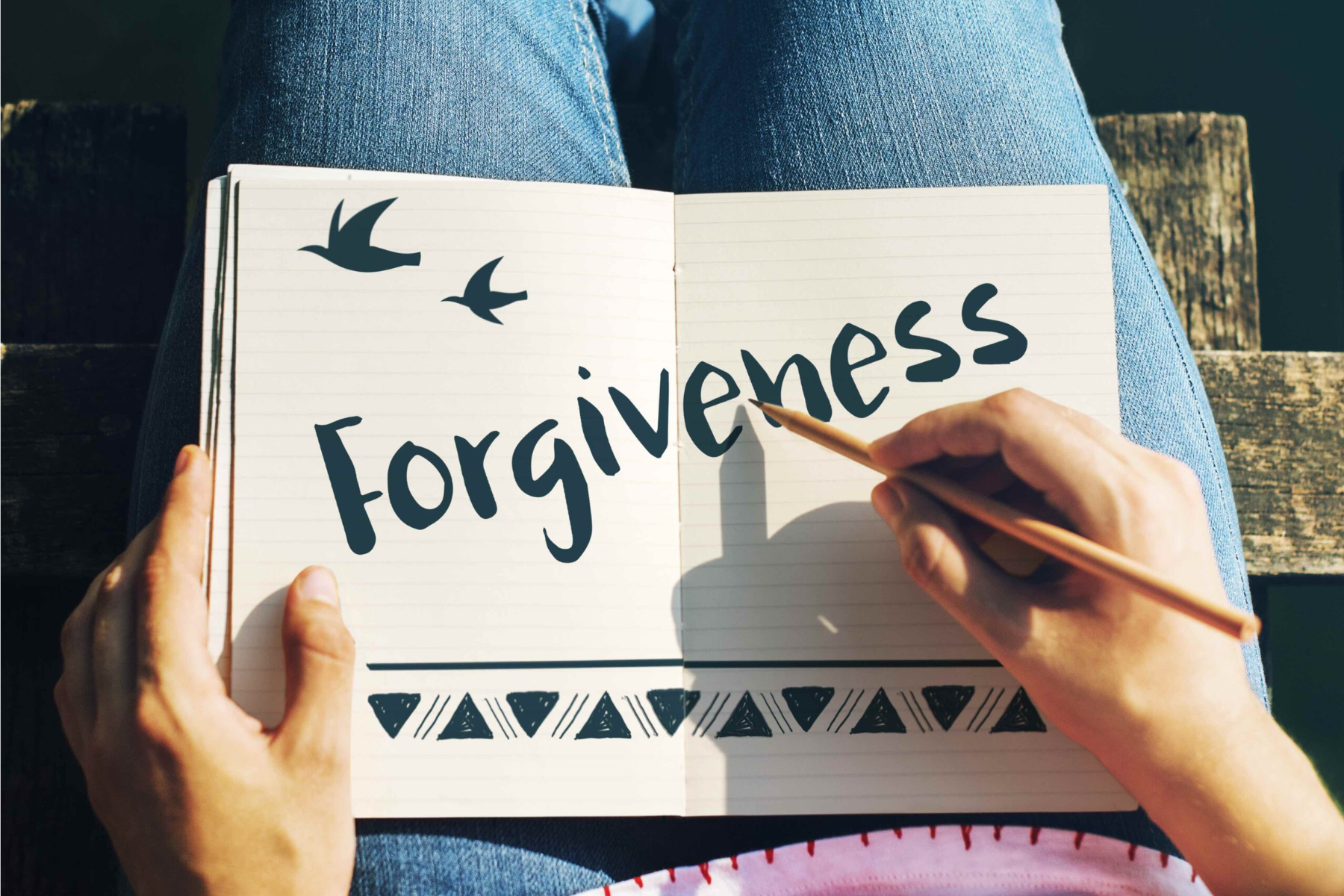 A hand holding a pen writes the word "Forgiveness" on a notebook page