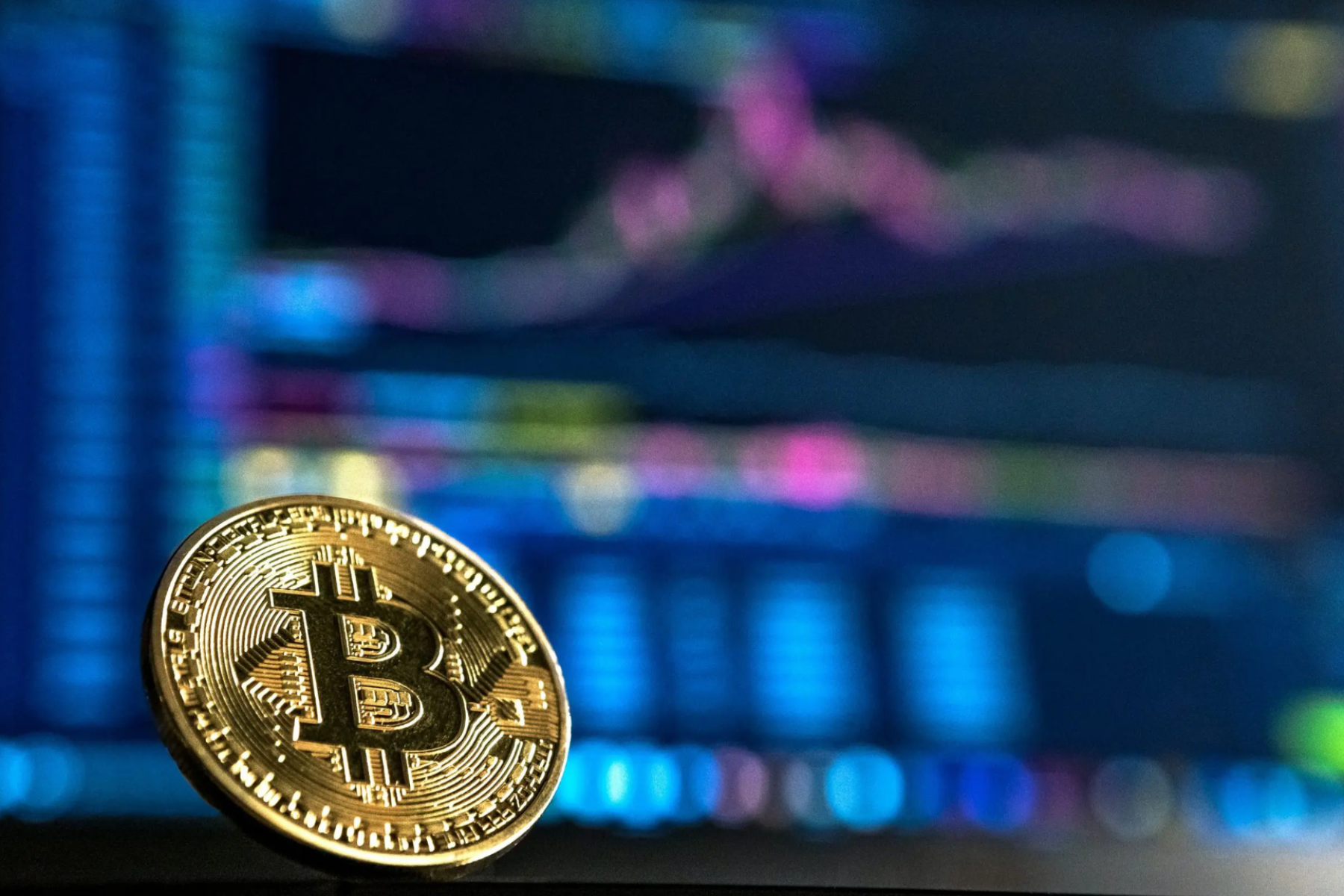 A single golden Bitcoin is placed in front of a computer screen with a blurred background
